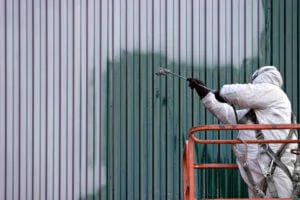 A painting contractor in protective suit uses a spray gun to paint a corrugated metal wall, standing on an orange lift.