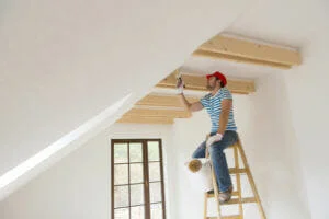 A man on a ladder providing a residential painting service for the ceiling of a room, wearing casual attire and a red cap. Natural light streams in through a window.
