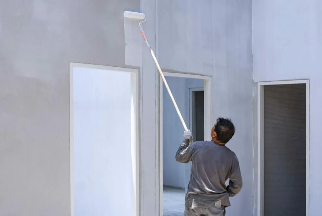 A painting contractor using a long-handled roller to paint an exterior wall white.