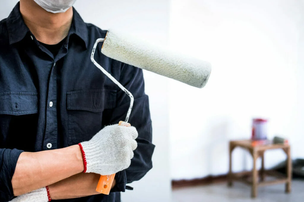 A professional painter in Central Florida in a mask and gloves holding a paint roller, with a partially painted wall and a paint tray in the background.